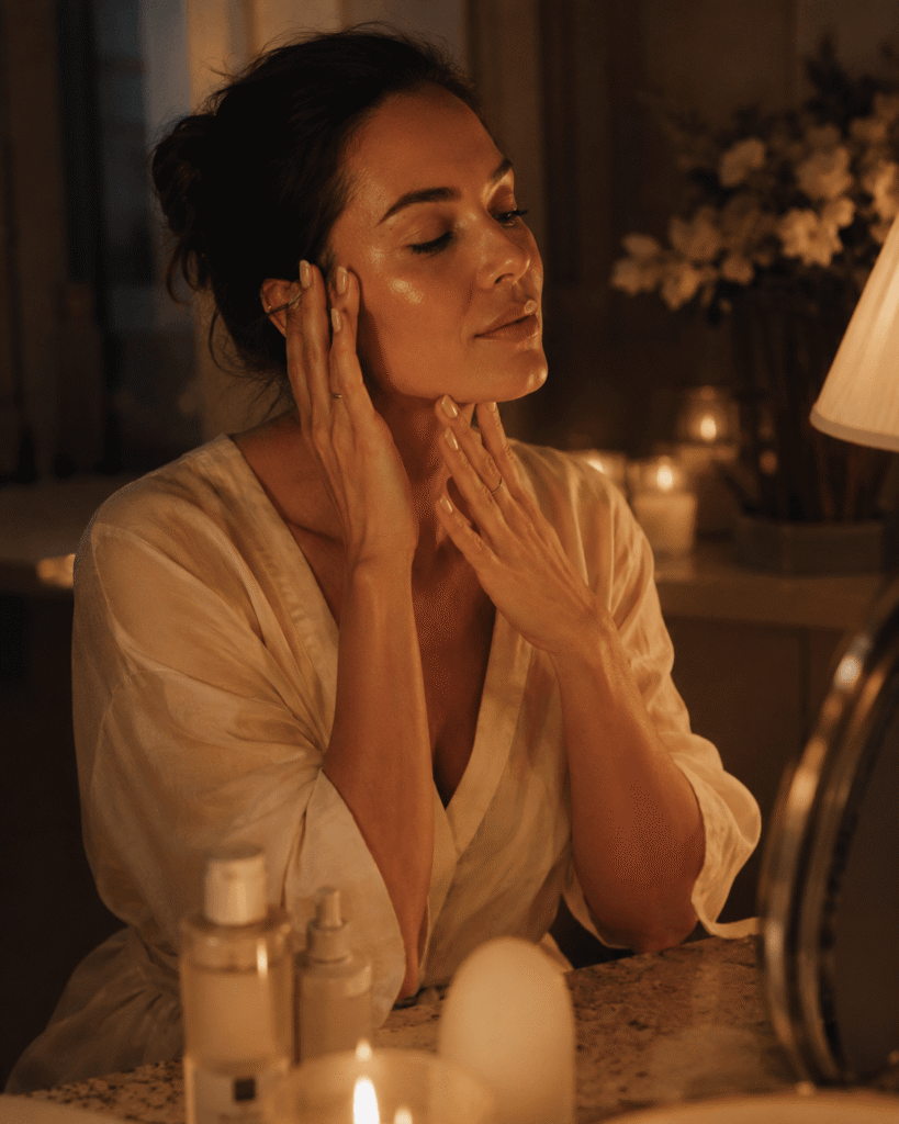 Woman in a cream robe gently massaging her face at a vanity with warm candlelight and skincare products nearby.