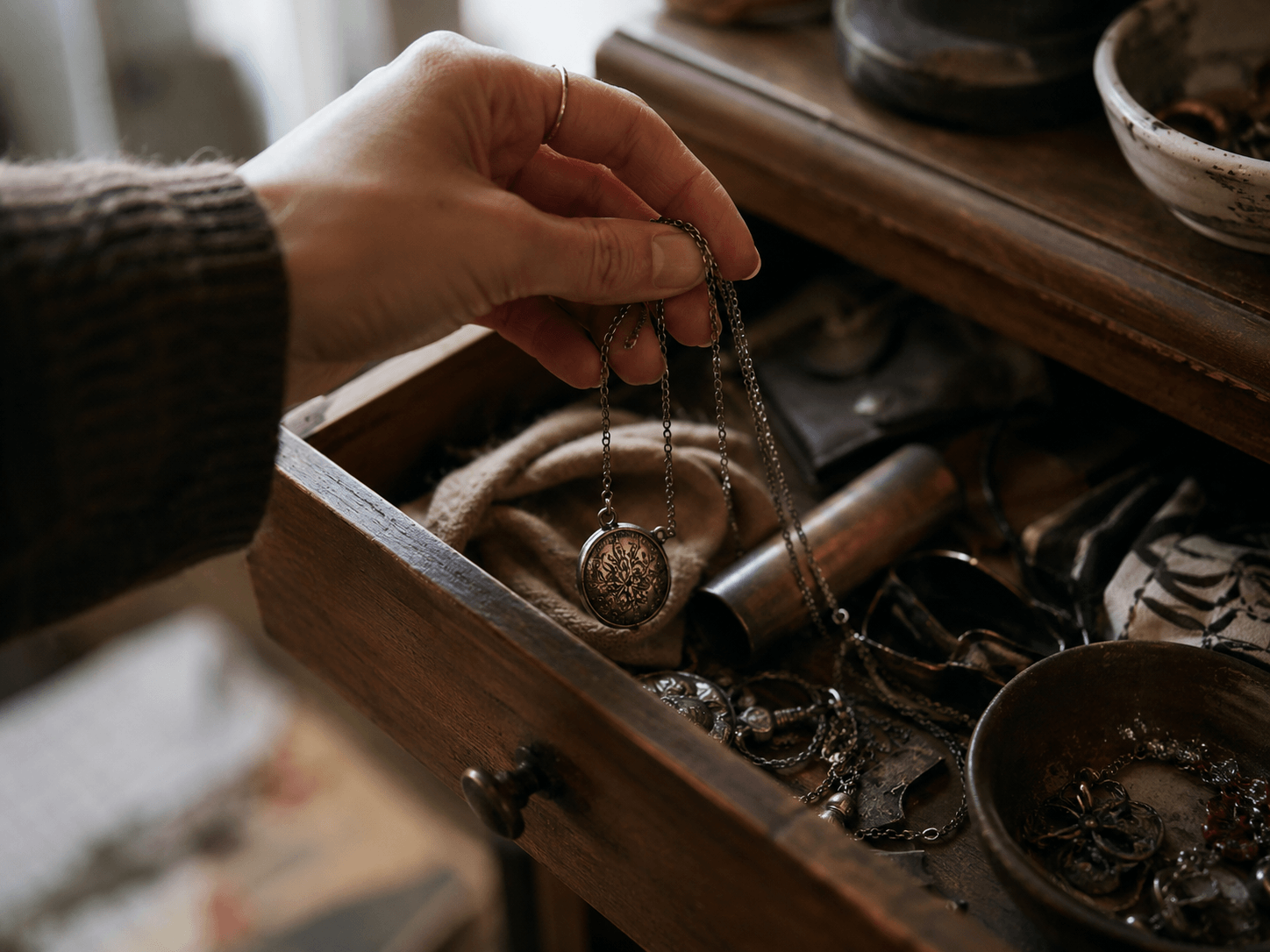 A woman’s hand pulling a tarnished silver necklace from a cluttered wooden drawer in soft natural light.