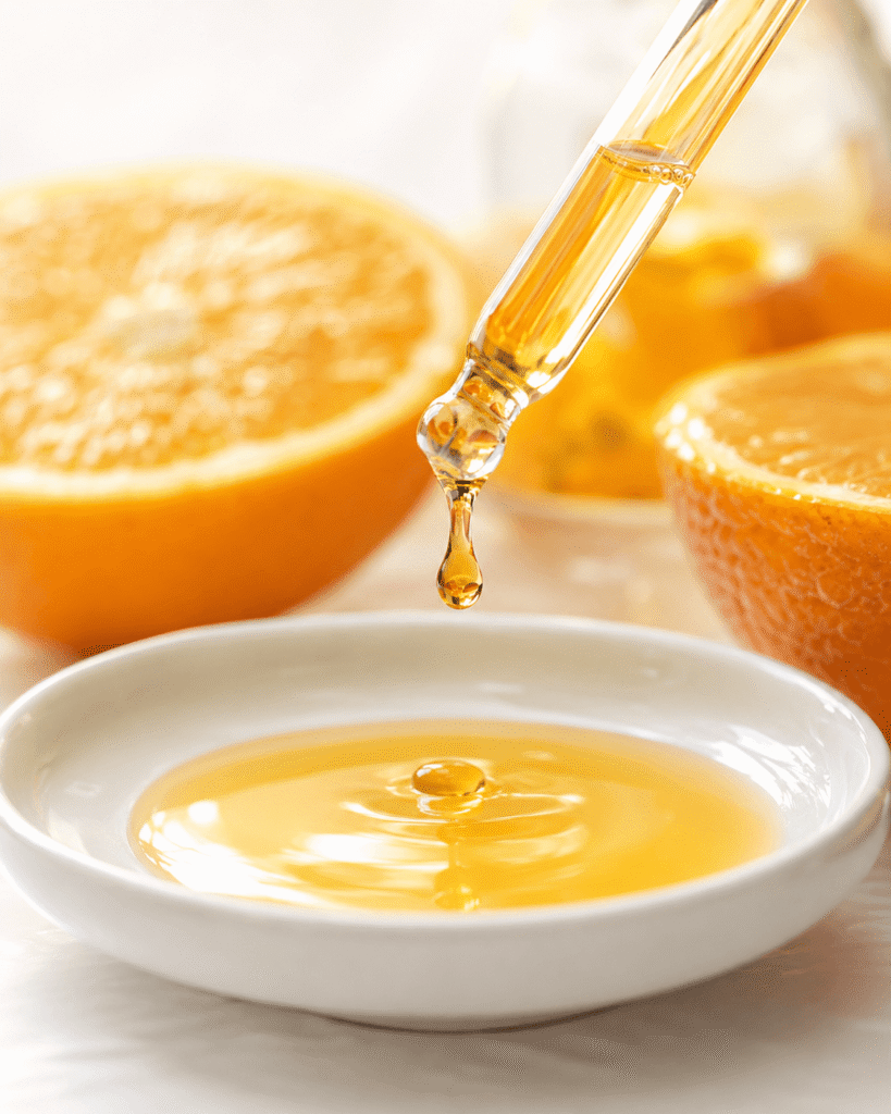 Macro photograph of golden orange serum dripping from a glass dropper into a white ceramic dish with blurred orange halves behind.